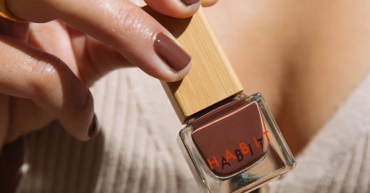 Hand holding a bottle of Habit brown nail polish with a blurred background